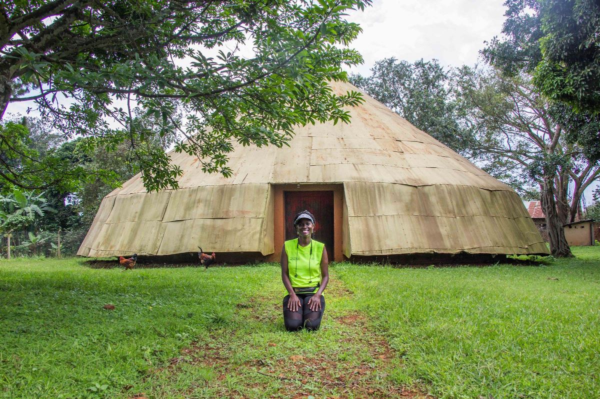 A photograph of a tourist kneeling infront of the Buganda House at Naggalabi Buddo, Uganda