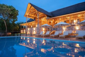 Stunning evening glow at Pabidi Lodge’s swimming pool, reflecting the elegant thatched-roof main lodge nestled in the forest