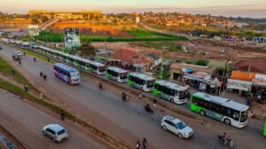 An aerial view shows a long line of Kayoola Electric Buses moving through a busy Kampala-area road at golden hour, marking the arrival of the new zero-emission fleet for routes serving Kira Municipality and surrounding communities