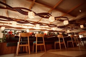 The spacious dining hall at Café Shires, highlighted by undulating wooden ceiling fixtures, cube pendant lights, and booth seating lined with vibrant plants and flowers