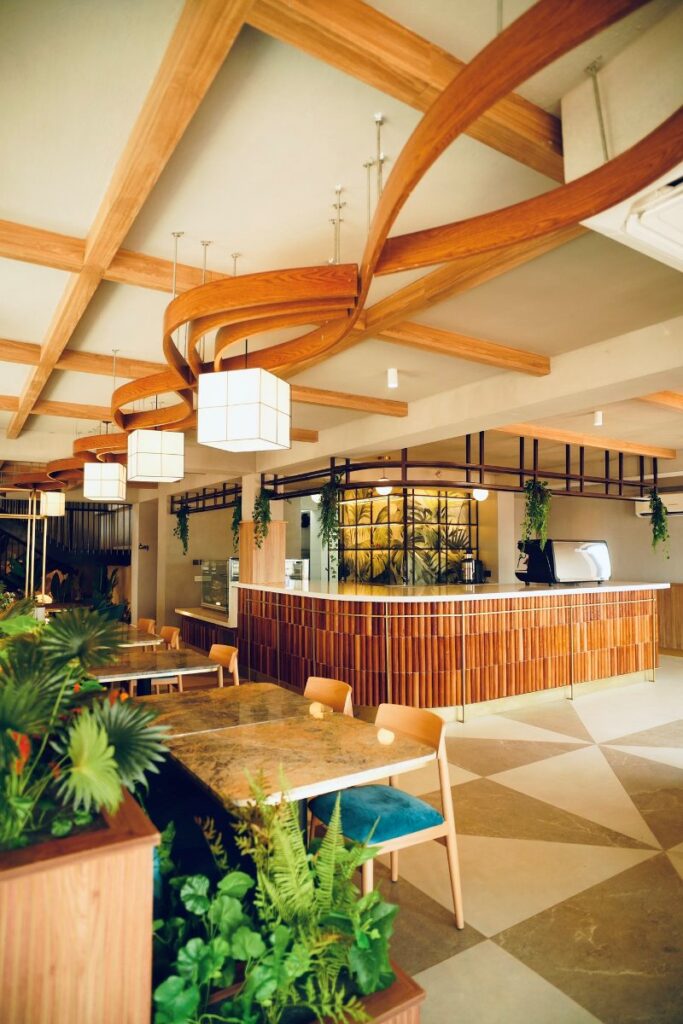 The main dining area and service counter at Café Shires in Tororo, featuring a sculptural curved wooden ceiling, hanging cube pendant lights, and abundant indoor greenery
