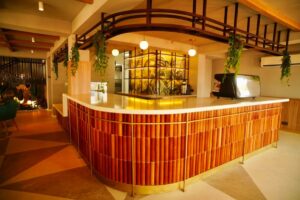 The distinctive curved bar at Café Shires, clad in vertical wooden rods with a white countertop, espresso machine, and illuminated tropical leaf mural in the background